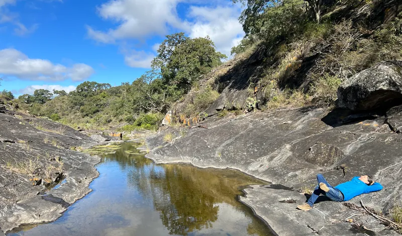 Una persona descansa recostada sobre las rocas lisas, a orillas del río, entre el bosque y la sierra